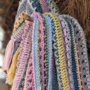 May include: A colorful, crocheted scarf in pastel shades of pink, blue, yellow, and gray. The scarf features a variety of stitch patterns and a tassel. The scarf is draped over a small branch, showcasing its texture and design.