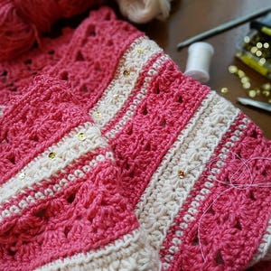 Könnte beinhalten: Eine rosa und weiße Häkeldecke mit goldenen Pailletten. Die Decke ist in der Mitte gefaltet und hat einen weißen Häkelrand.
