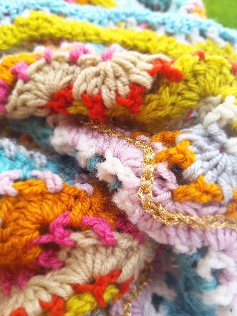 May include: Close-up of a colourful crocheted blanket with a gold chain detail. The blanket is made with a variety of different coloured yarns, including pink, blue, yellow, orange, and white.