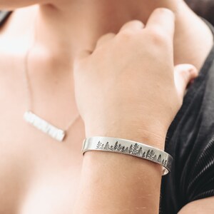 May include: A silver bracelet with a tree silhouette design. The bracelet is worn on a person's wrist.