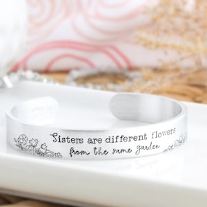 May include: Silver bracelet with the inscription "Sisters are different flowers from the same garden".