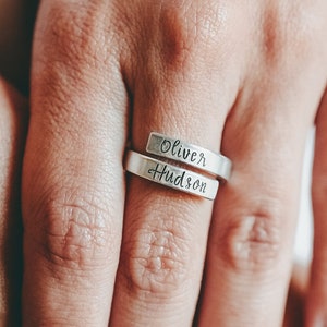 May include: A silver ring with the names "Oliver" and "Hudson" engraved on it. The ring is worn on a person's finger.
