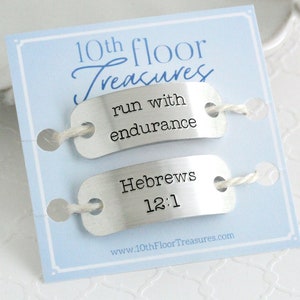 May include: A light blue card with the text "10th floor Treasures" and two silver tags. The top tag reads "run with endurance" and the bottom tag reads "Hebrews 12:1". The tags are connected with white string.