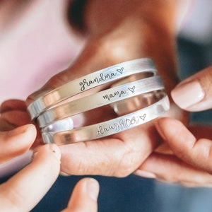 May include: Three silver bangle bracelets with the words "grandma", "mama", and "daughter" engraved on them, each with a small heart symbol.
