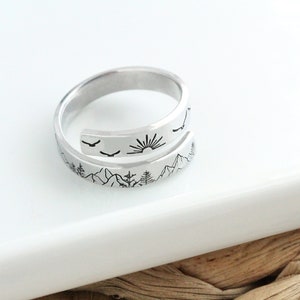 May include: A silver ring with a mountain, sun, and birds design. The ring is open and adjustable.