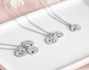May include: Three silver necklaces with small round pendants. Each pendant has a different letter and a small gemstone. The gemstones are blue, green, and red.