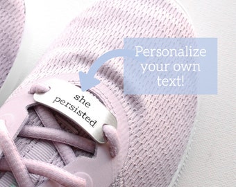 May include: A pair of pink running shoes with a silver shoe charm that says "she persisted". The charm is attached to the laces of the shoe. The text "Personalize your own text!" is written on a blue rectangle above the shoe.