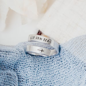 May include: A silver ring with the words "Let Him Lead" engraved on it. A small cross is engraved on the other side of the ring.