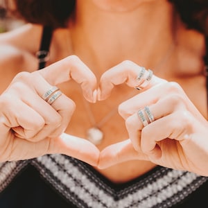 May include: A person wearing a black and white striped top makes a heart shape with their hands. They are wearing several silver rings with engraved words, including "GRIT" and "a prayer".