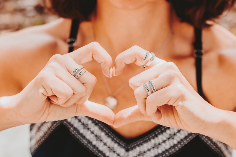 May include: A person wearing a black and white striped top makes a heart shape with their hands. They are wearing several silver rings with engraved words, including "GRIT" and "a prayer".
