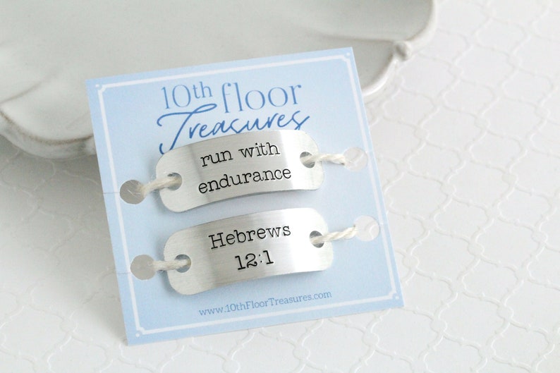May include: A light blue card with the text "10th floor Treasures" and two silver tags. The top tag reads "run with endurance" and the bottom tag reads "Hebrews 12:1". The tags are connected with white string.