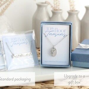 May include: Two options for packaging a silver necklace with a dandelion design. The first option is a clear plastic bag with a white card that says "10th Floor Treasures". The second option is a blue gift box with a white ribbon and a white card that says "10th Floor Treasures".