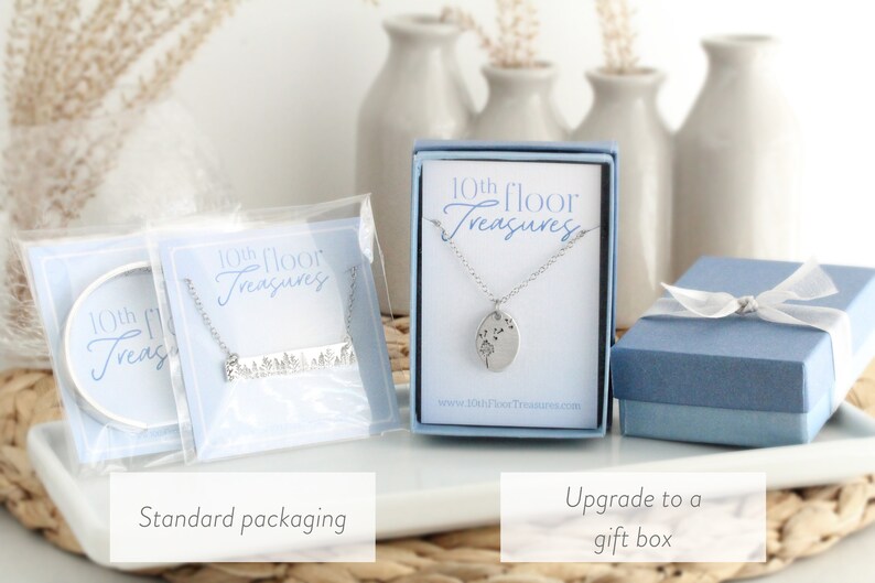 May include: Two options for packaging a silver necklace with a dandelion design. The first option is a clear plastic bag with a white card that says "10th Floor Treasures". The second option is a blue gift box with a white ribbon and a white card that says "10th Floor Treasures".