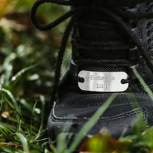 Puede incluir: Primer plano de una zapatilla deportiva negra con una etiqueta plateada. La etiqueta lleva la inscripci&oacute;n "Hebreos 12:1". La zapatilla est&aacute; atada con cordones negros y se encuentra en hierba verde.