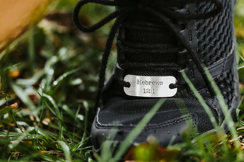 Puede incluir: Primer plano de una zapatilla deportiva negra con una etiqueta plateada. La etiqueta lleva la inscripci&oacute;n "Hebreos 12:1". La zapatilla est&aacute; atada con cordones negros y se encuentra en hierba verde.