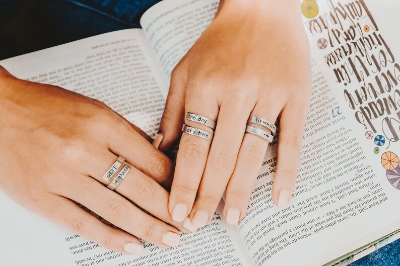 May include: Close-up of a person's hands wearing three silver rings with engraved messages. The rings say "one day at a time", "till we meet again", and "grit & grace".