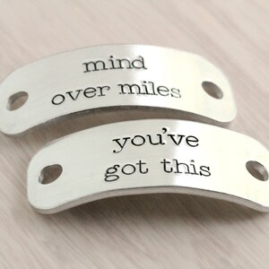 Puede incluir: Dos abalorios de zapatos plateados con mensajes inspiradores. El abalorio superior dice "mind over miles" y el abalorio inferior dice "you've got this".