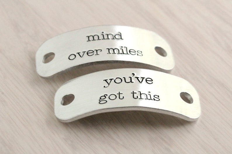 Puede incluir: Dos abalorios de zapatos plateados con mensajes inspiradores. El abalorio superior dice "mind over miles" y el abalorio inferior dice "you've got this".