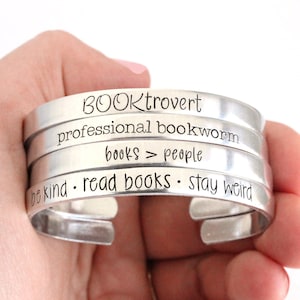 May include: Silver bracelet with four stacked bands. The bands are engraved with the phrases "BOOKtrovert", "professional bookworm", "books > people", and "be kind - read books - stay weird".