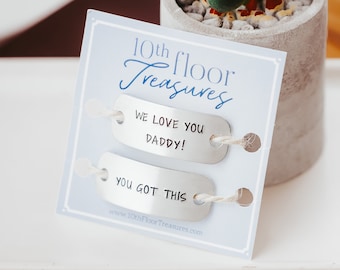 May include: Two silver metal tags with the text "We love you Daddy!" and "You got this" on a white card with the text "10th floor Treasures" and a website address.