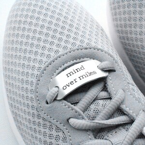 Puede incluir: Zapatillas de correr grises con una etiqueta de metal plateado que dice "mind over miles" en los cordones.