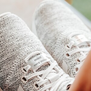 Puede incluir: Zapatillas blancas para correr con una etiqueta de metal plateado que dice "mind over miles".