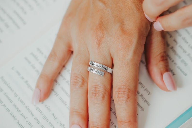 May include: Silver wrap ring with the words "just breathe" engraved on the band. The ring also features a small floral design. The ring is on a finger, resting on a page of a book.