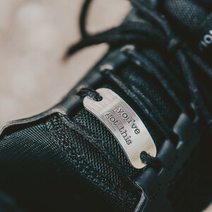 Puede incluir: Primer plano de un zapato negro con una etiqueta de metal plateado que dice "You've got this".