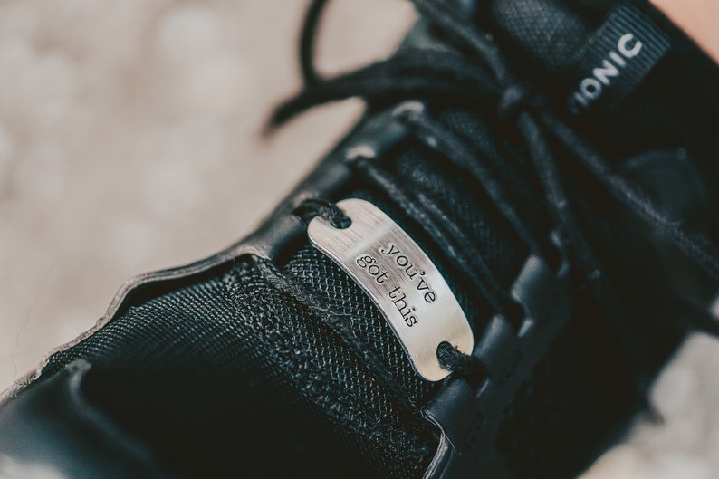 Puede incluir: Primer plano de un zapato negro con una etiqueta de metal plateado que dice "You've got this".