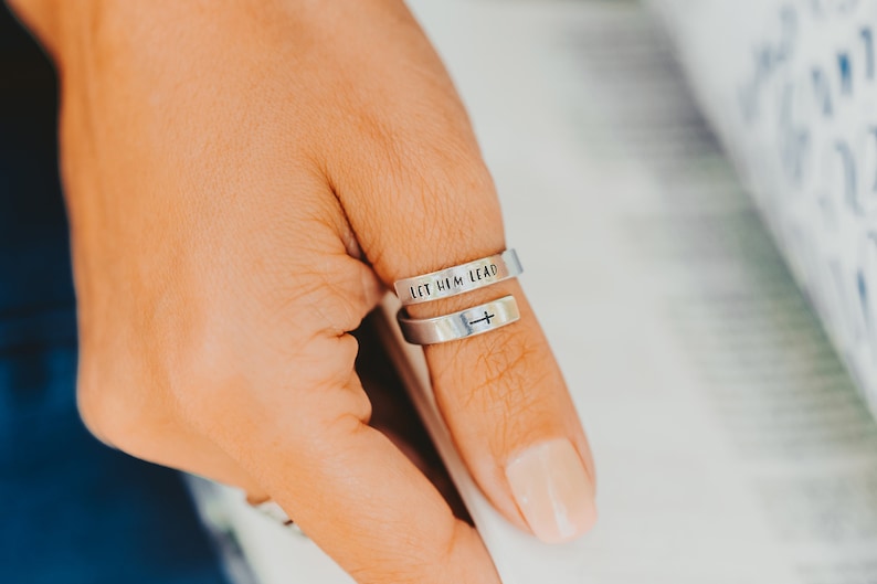 May include: A silver ring with the words "Let Him Lead" engraved on one band and a cross on the other band.