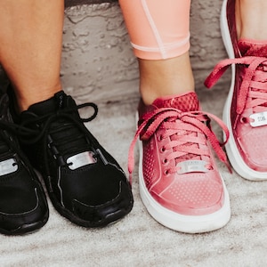 Puede incluir: Dos pares de zapatillas deportivas, unas negras y otras rosas, con suelas blancas. Cada zapatilla tiene una etiqueta met&aacute;lica en los cordones. Las zapatillas negras tienen el texto "Bed with Breakfast" y las rosas "Yummy Vision" y "Dom - Run".