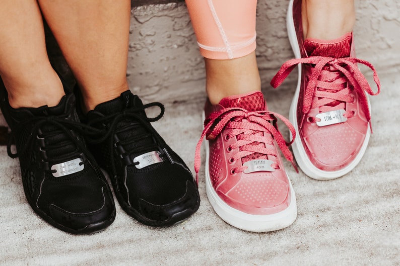 Puede incluir: Dos pares de zapatillas deportivas, unas negras y otras rosas, con suelas blancas. Cada zapatilla tiene una etiqueta met&aacute;lica en los cordones. Las zapatillas negras tienen el texto "Bed with Breakfast" y las rosas "Yummy Vision" y "Dom - Run".