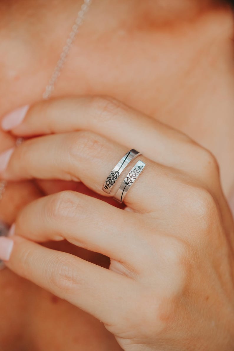 May include: A silver-colored wrap ring with floral engravings. The ring features a split design, with the floral details on the outer bands. The ring is worn on a finger, showcasing the delicate design and craftsmanship.