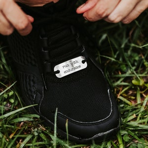 Puede incluir: Zapatilla deportiva negra con una etiqueta plateada que dice "run with endurance". La etiqueta es rectangular con bordes redondeados y dos agujeros para los cordones. La zapatilla est&aacute; sobre hierba verde.