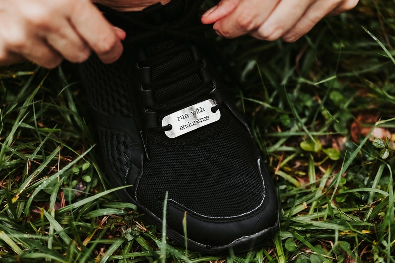 Puede incluir: Zapatilla deportiva negra con una etiqueta plateada que dice "run with endurance". La etiqueta es rectangular con bordes redondeados y dos agujeros para los cordones. La zapatilla est&aacute; sobre hierba verde.