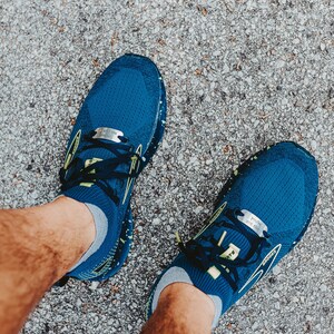 Puede incluir: Vista superior de zapatillas de correr azules con cordones negros y detalles amarillos. Las zapatillas est&aacute;n sobre una superficie gris texturizada. Las zapatillas tienen una etiqueta plateada en los cordones.