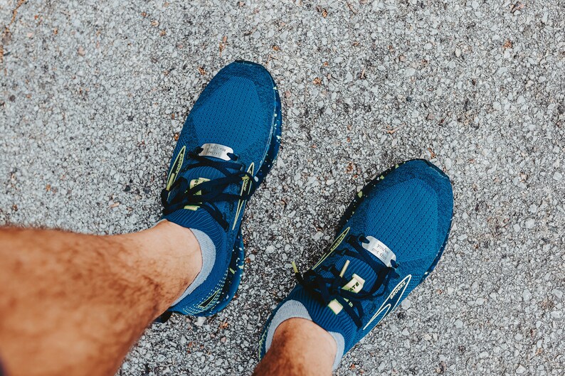 Puede incluir: Vista superior de zapatillas de correr azules con cordones negros y detalles amarillos. Las zapatillas est&aacute;n sobre una superficie gris texturizada. Las zapatillas tienen una etiqueta plateada en los cordones.