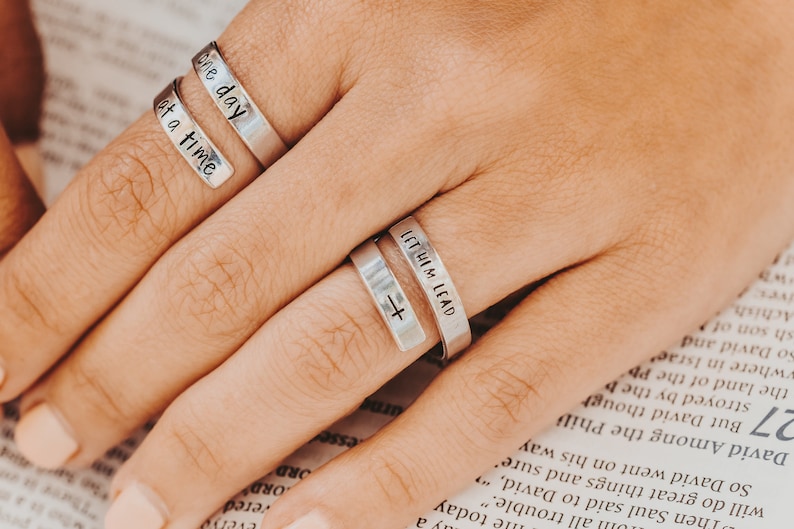 May include: Three silver rings with engraved messages. The top ring says "one day at a time". The middle ring says "let him lead" with a small cross symbol. The bottom ring is plain.