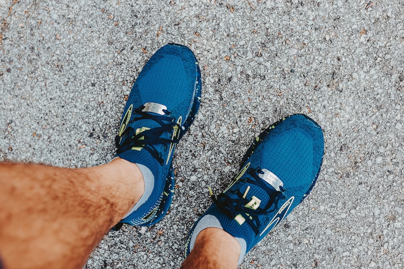 Puede incluir: Un par de zapatillas de running azules con cordones negros y una etiqueta plateada en cada zapato. Las zapatillas est&aacute;n sobre una superficie de grava gris.