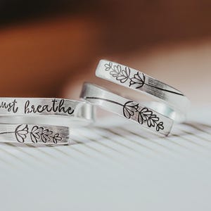 May include: Three silver rings with engraved designs. The top ring has a floral design, the middle ring has a floral design, and the bottom ring has the words "just breathe" engraved on it.