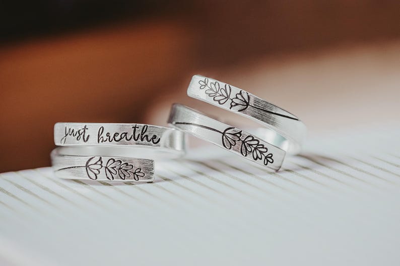 May include: Three silver rings with engraved designs. The top ring has a floral design, the middle ring has a floral design, and the bottom ring has the words "just breathe" engraved on it.