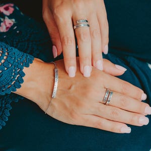 May include: A silver bracelet with a long, thin, engraved bar charm. The bracelet is worn on the wrist of a person wearing a blue floral dress. The person is also wearing two silver rings on their fingers.