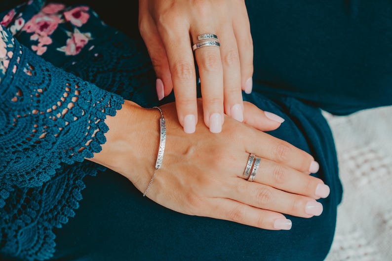 May include: A silver bracelet with a long, thin, engraved bar charm. The bracelet is worn on the wrist of a person wearing a blue floral dress. The person is also wearing two silver rings on their fingers.