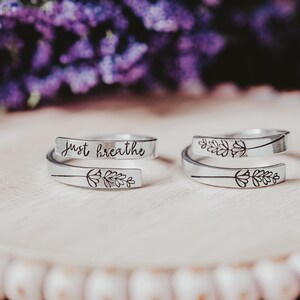 May include: Two silver-colored wrap rings with floral designs and the words "just breathe" engraved on one. The rings are displayed on a light-colored wooden surface, with blurred purple flowers in the background.