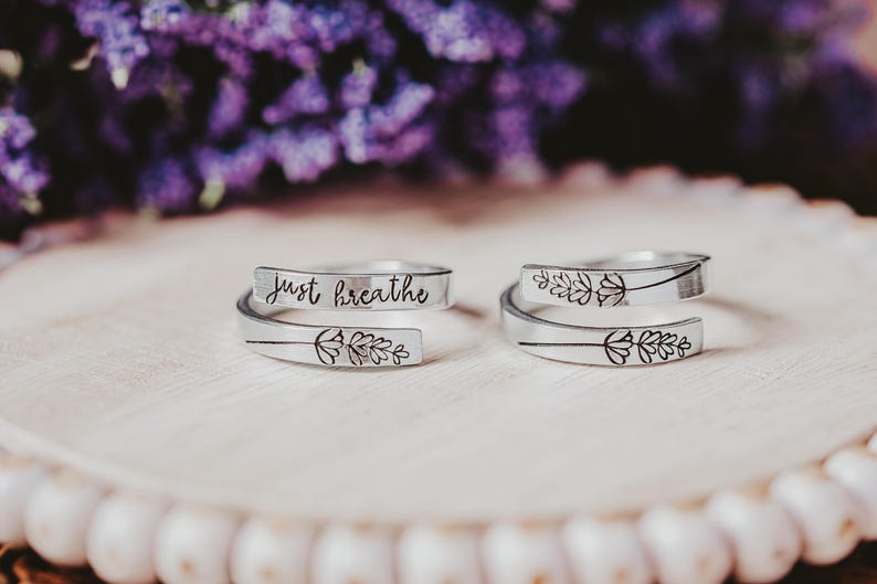 May include: Two silver-colored wrap rings with floral designs and the words "just breathe" engraved on one. The rings are displayed on a light-colored wooden surface, with blurred purple flowers in the background.
