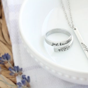 May include: A silver ring with the words "Just breathe" engraved on it and a silver necklace with a bar pendant featuring a simple flower design.