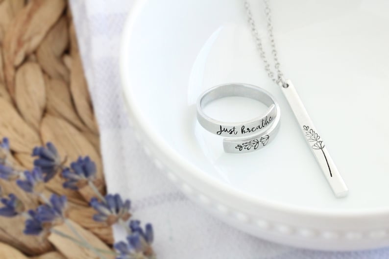May include: A silver ring with the words "Just breathe" engraved on it and a silver necklace with a bar pendant featuring a simple flower design.