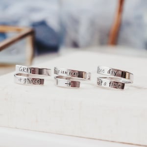 May include: Three silver rings with inspirational messages engraved on them. The rings are stacked on a white surface. The messages on the rings are "GRIT & grace", "LET HIM LEAD" with a cross symbol, and "one day at a time".