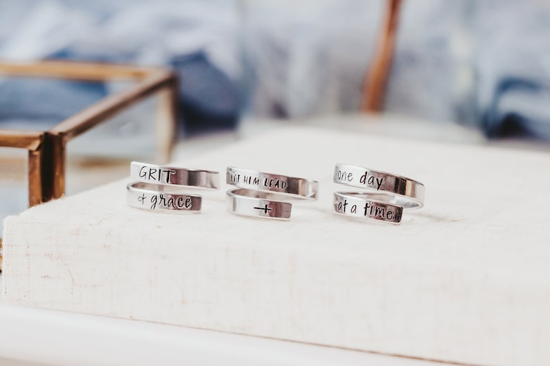 May include: Three silver rings with inspirational messages engraved on them. The rings are stacked on a white surface. The messages on the rings are "GRIT & grace", "LET HIM LEAD" with a cross symbol, and "one day at a time".