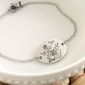 May include: A silver chain bracelet with a round charm featuring a black and white illustration of a book with flowers and butterflies.